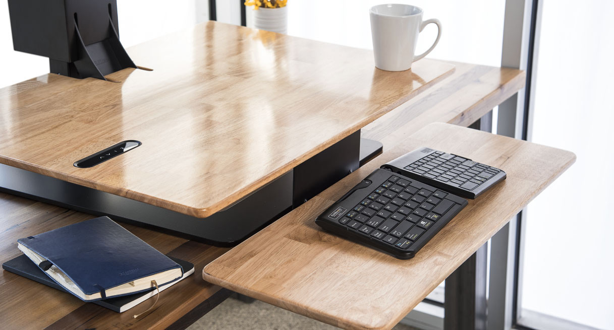 UPLIFT E7 electric desk converter with a wood-look surface and a lower keyboard tray holding a keyboard